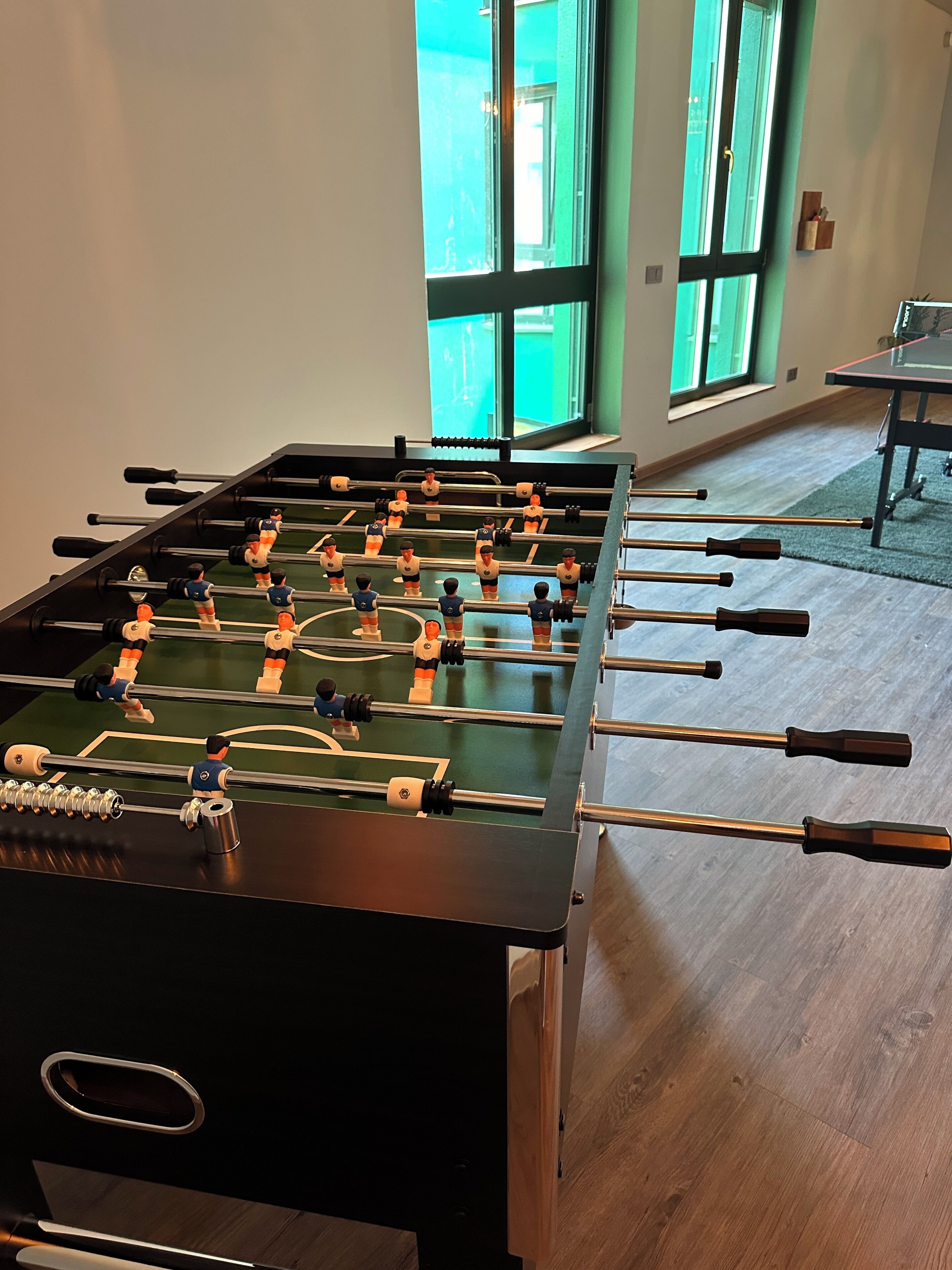 Una mesa de fútbol de mesa en una habitación con suelo de madera y ventanas grandes.