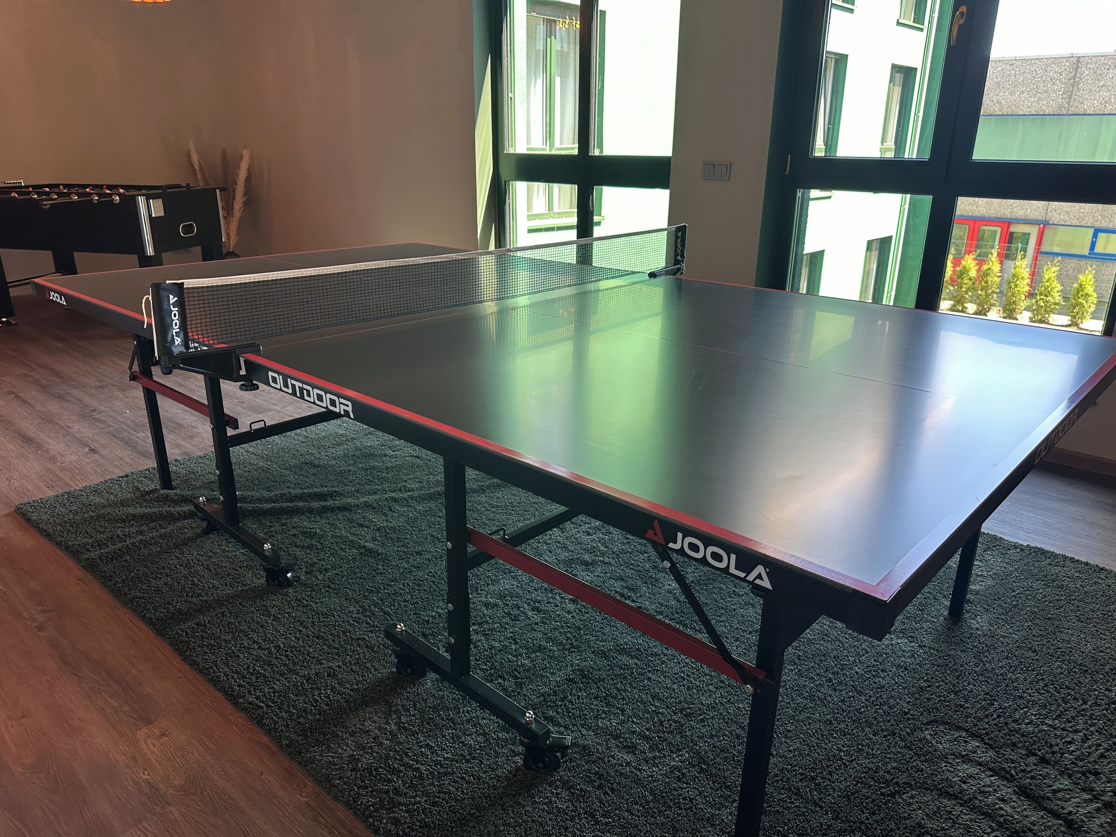 A ping pong table with "Joola" branding, positioned on a carpeted floor with a wooden table nearby.