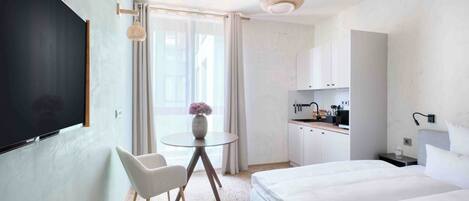 Een moderne hotelkamer met een bed, een bureau met stoel, een klein tafeltje met vaas en een kitchenette.