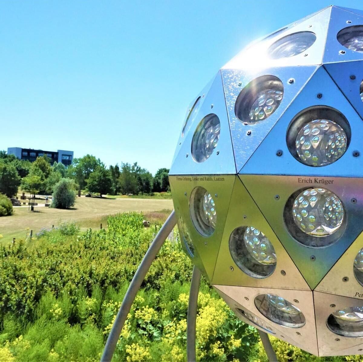 Una escultura esférica metálica con superficies reflectantes e inscripciones de texto, sobre un fondo de vegetación y cielo despejado.