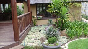 Une construction à toit de chaume dotée d’une terrasse en bois, entourée de différentes plantes et d’un petit jardin.