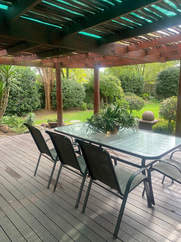 Un patio techado al aire libre con una mesa y sillas de vidrio, rodeado de abundante vegetación y árboles.