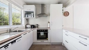 Une cuisine compacte avec des meubles blancs, un four encastré, une machine à café et une fenêtre équipée de stores.