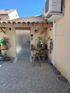 A small outdoor area with a metal door, potted plants, a stool, and a small table with a mirror and decorative items.