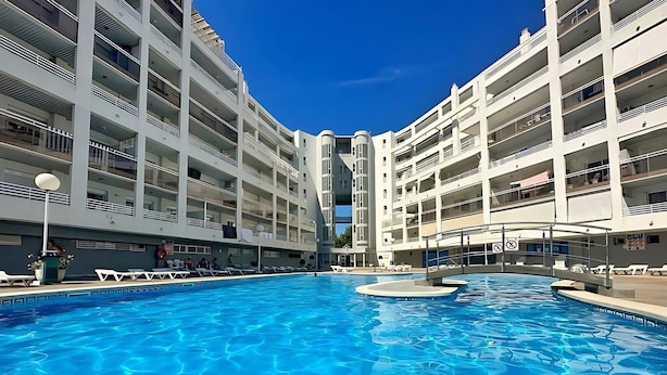 A modern hotel with a central swimming pool, multiple balconies, and a clear blue sky.