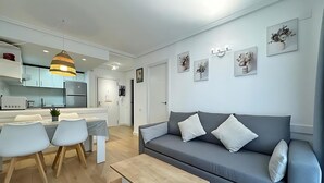 A modern living room with a grey sofa, a dining table with chairs, and a kitchen area in the background.
