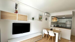 A modern living room with a flat-screen TV, a wooden bench, and a dining area with chairs and a table.