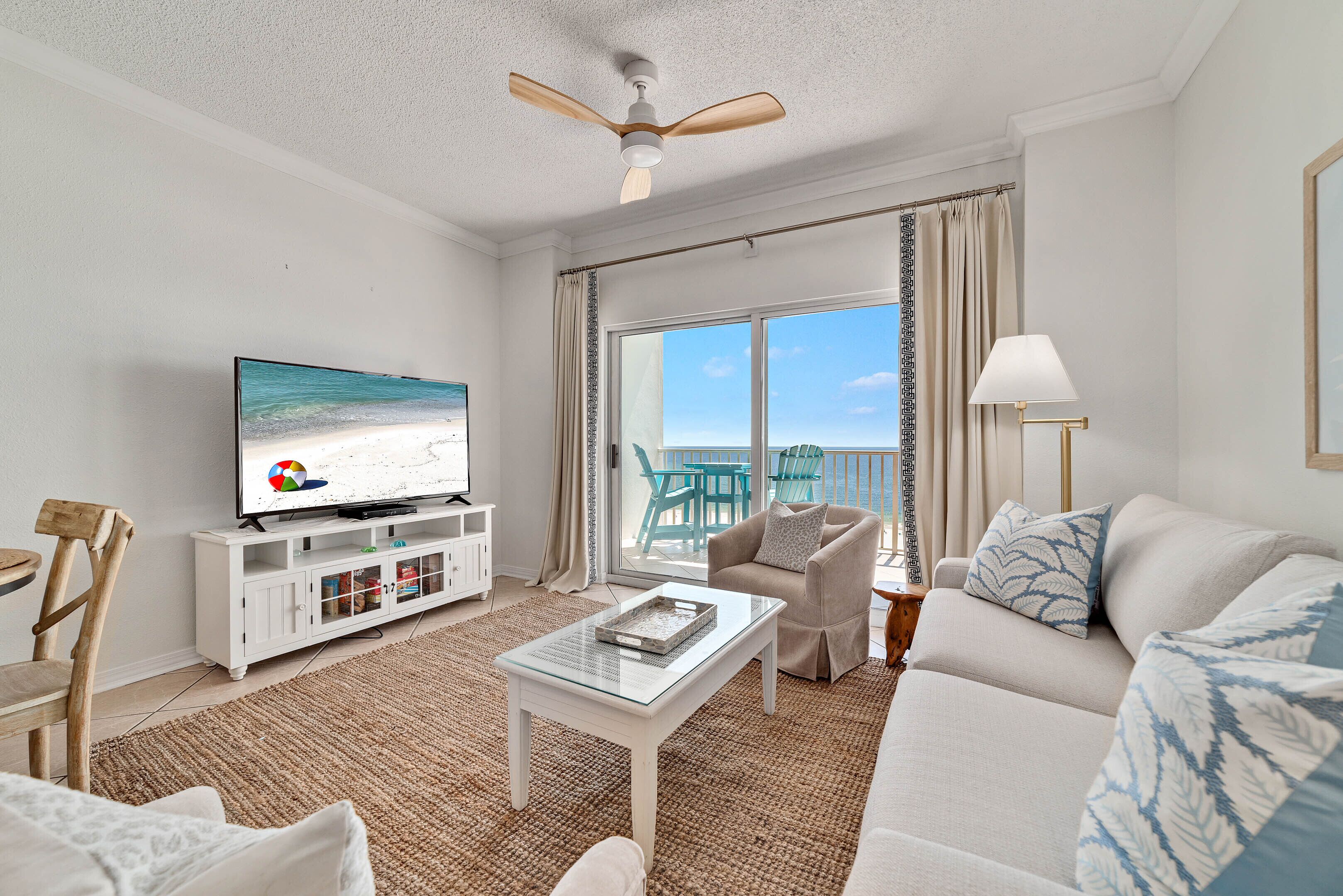 Ein Wohnzimmer mit Fernseher, weißen Möbeln, Deckenventilator und Blick ins Freie.