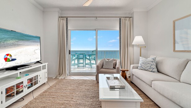 A living room with a television, a sofa, a coffee table, and a view of the beach through the sliding glass door. - Tidewater 907-Family friendly complex to enjoy the Beach! (Orange Beach)
