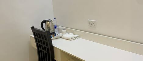 Una habitación pequeña y limpia con un escritorio blanco, una silla negra, un hervidor de agua, una botella de agua y una taza.