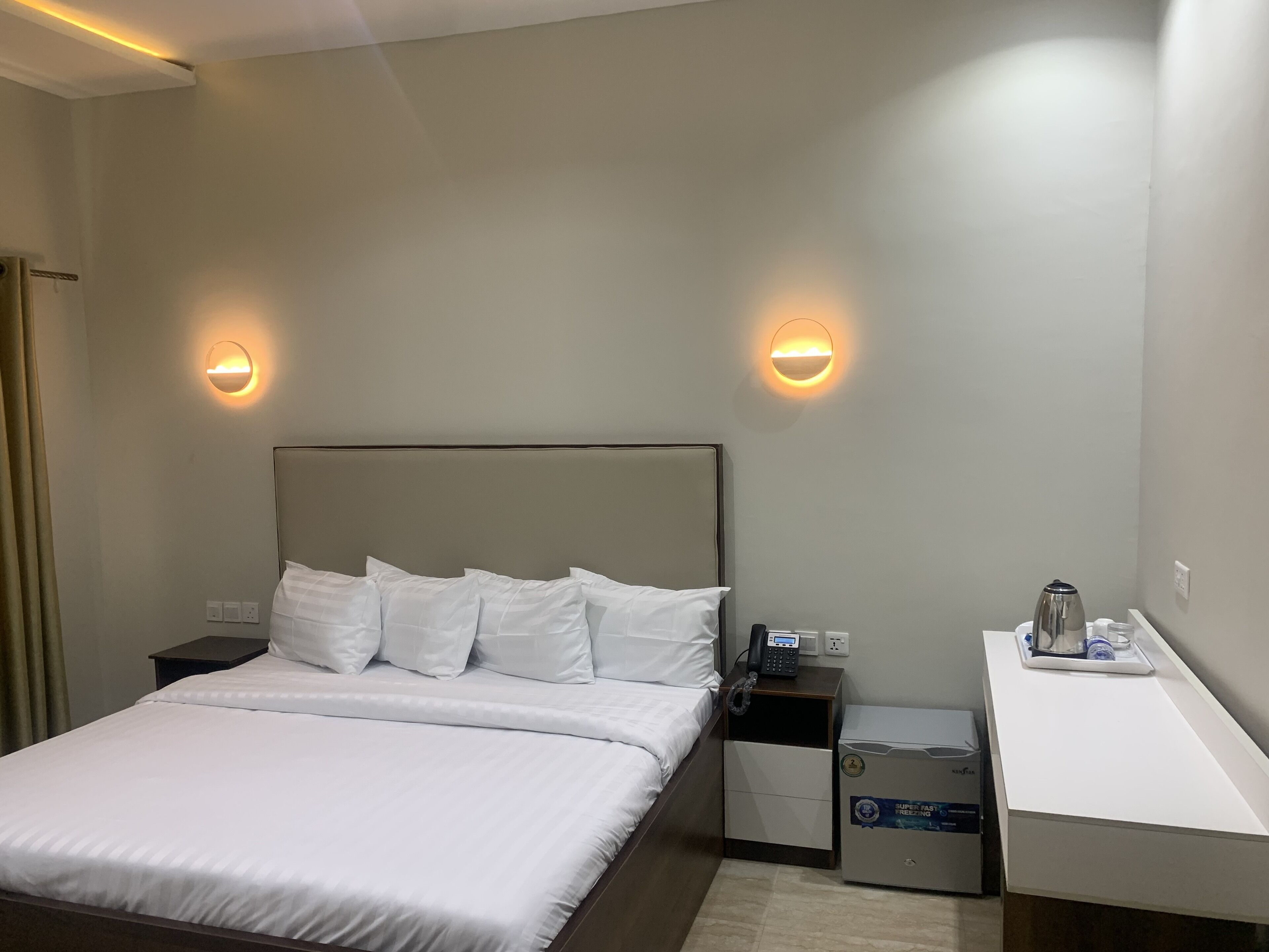 A hotel room with a bed, bedside tables, a desk with a kettle, and wall-mounted lights.