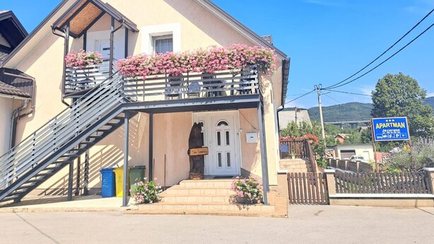A two-story building with a balcony adorned with pink flowers, a staircase leading to the entrance, and a sign indicating it is an apartment. - Apartments Jezerane (Brinje)