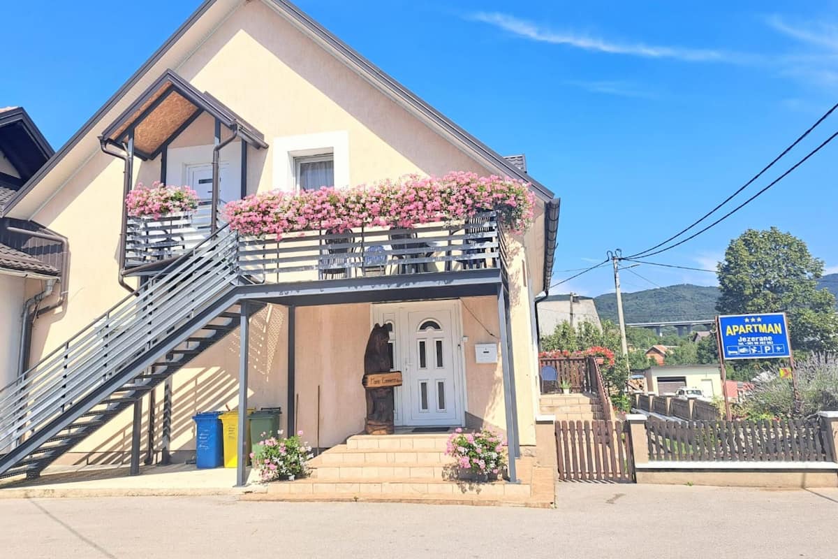 A two-story building with a balcony adorned with pink flowers, a staircase leading to the entrance, and a sign indicating it is an apartment.