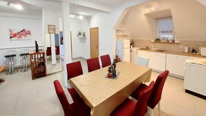 A modern kitchen with white cabinets and a wooden countertop. There is a dining area with a table and red chairs. The room has white walls and a tiled floor. - Apartments Jezerane (Brinje)