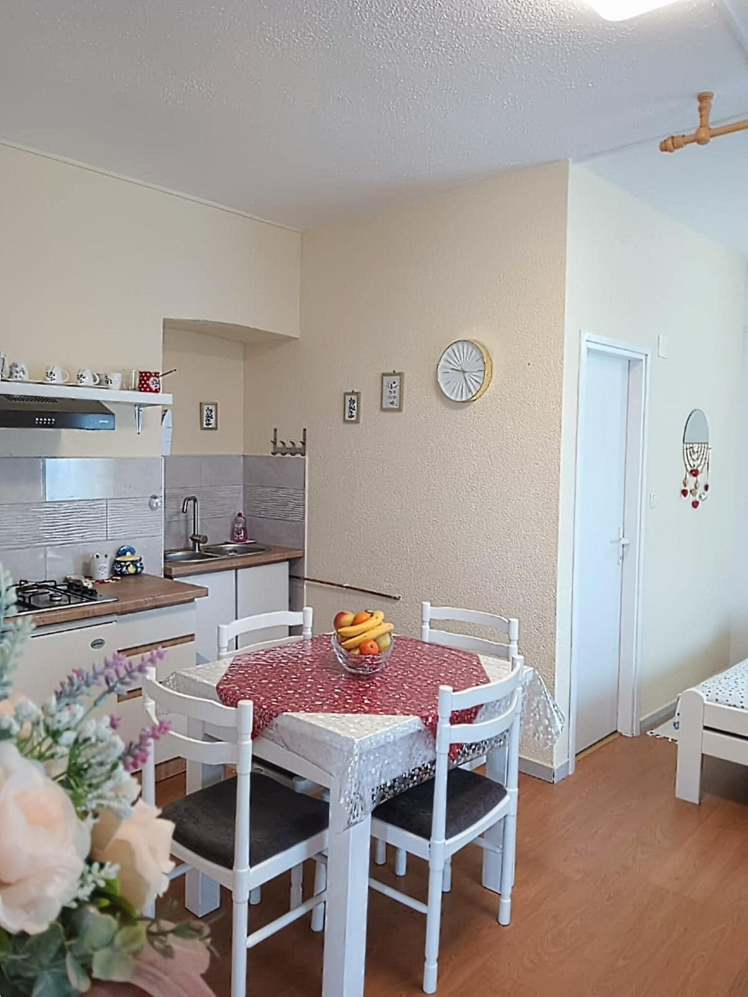A compact kitchen with a gas stove, a microwave, and a sink. There is a dining table with chairs and a bowl of fruit. The walls are plain with a clock and a dreamcatcher.
