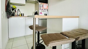 Een compacte keuken met witte kastjes, een magnetron en een koelkast. Er staan twee krukken met bruine kussens bij.
