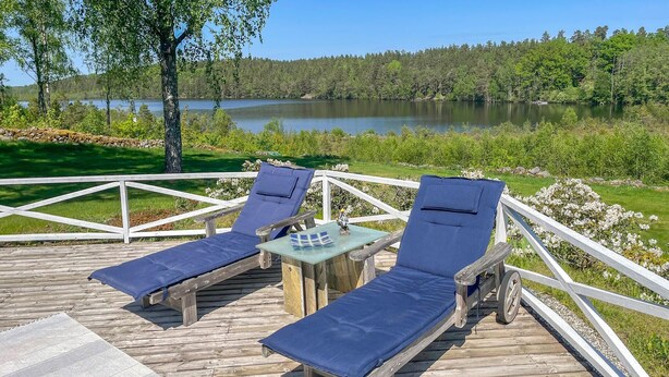 A wooden deck with two lounge chairs and a small table, overlooking a lake and forest. - Lovely home in Söderköping with sauna (Söderköping)