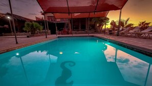 Una piscina con superficie de agua azul cristalina, rodeada de sillas de descanso y sombrillas, con un atardecer de fondo.