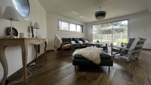 A spacious living room with a wooden floor, a sofa set, a rocking chair, a dining table, and a ceiling fan. - La Villa Case-bi, Profitez en Famille de ce Magnifique Logement (Saint-Germain-sur-Ay)