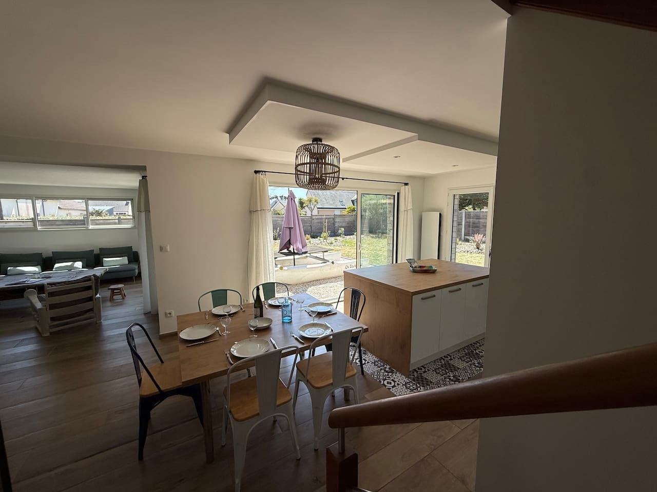 Een eetgedeelte met een houten tafel en stoelen, een keuken met een aanrecht, en een uitzicht op de omgeving door grote ramen.