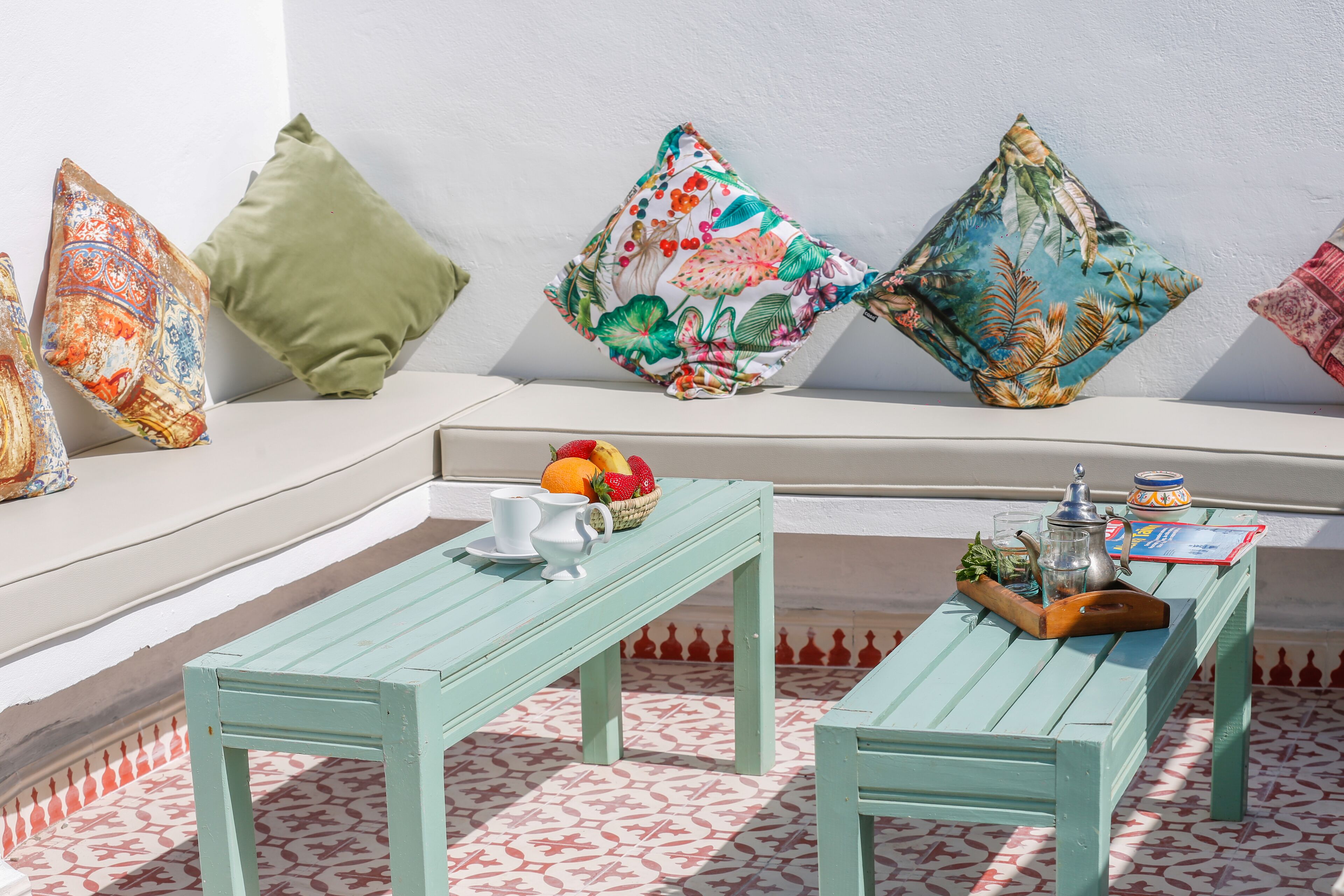 A sunlit outdoor seating area with a patterned floor, a wooden table, and colorful cushions.