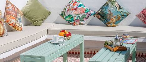 A sunlit outdoor seating area with a patterned floor, a wooden table, and colorful cushions.