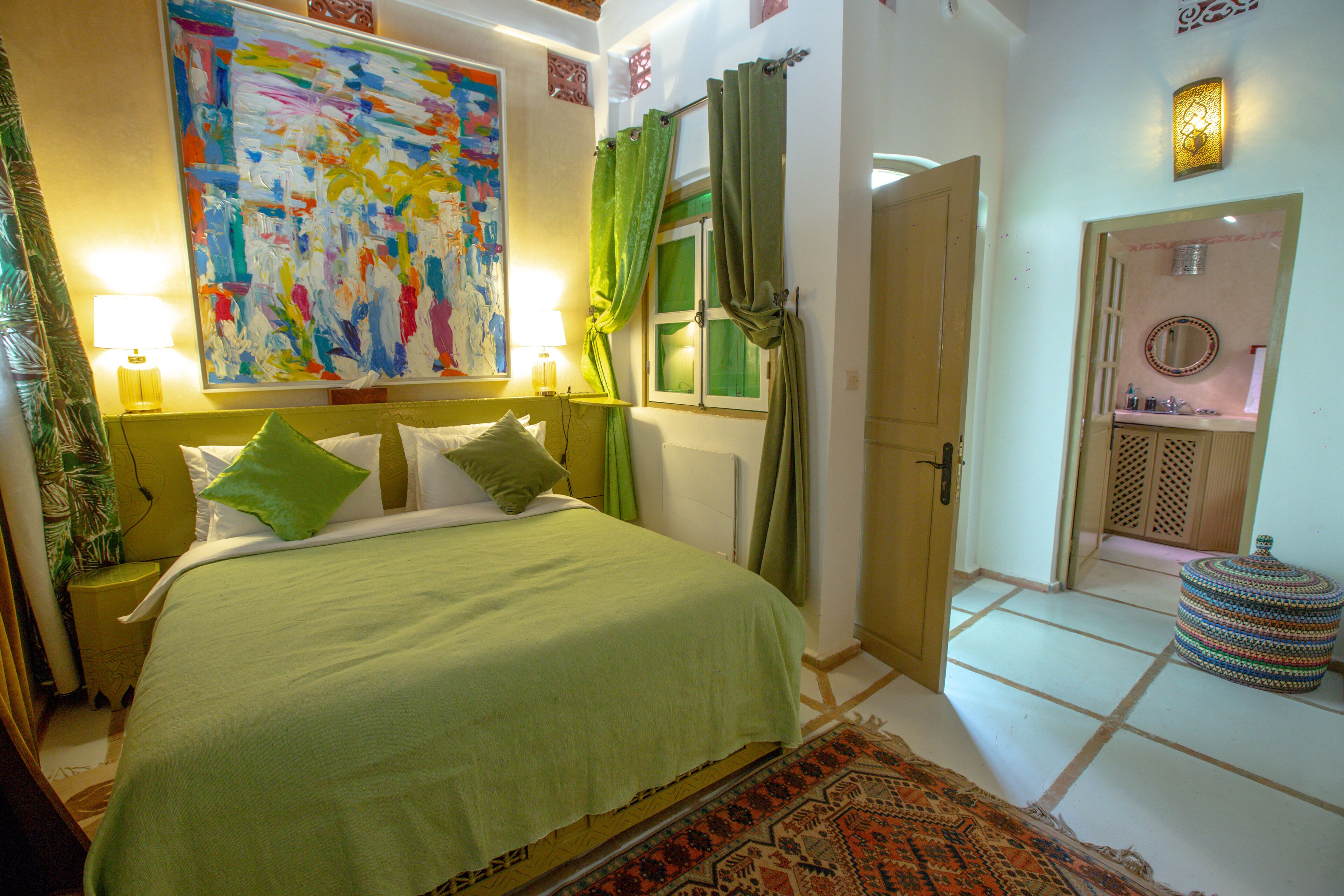 Un dormitorio con cama, almohadas, una mesita de noche, un cuadro, una lámpara, una puerta y una pequeña mesa con espejo.