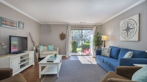 Sala de estar con televisor, sofá y mesa de centro. Cuenta con una puerta corrediza de vidrio que da a un balcón con mobiliario exterior.