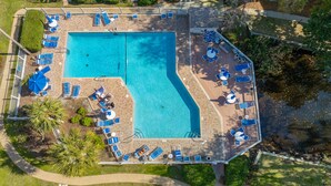 Vista aérea de una piscina rectangular con múltiples tumbonas y sombrillas, rodeada por un solárium empedrado y vegetación.