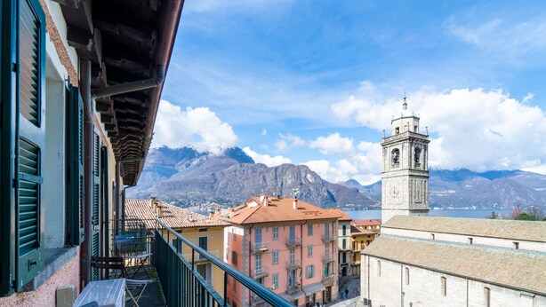 A view of a town with a prominent clock tower, colorful buildings, and mountains in the background. - Lotus Apartment With Lakeview (Bellagio)