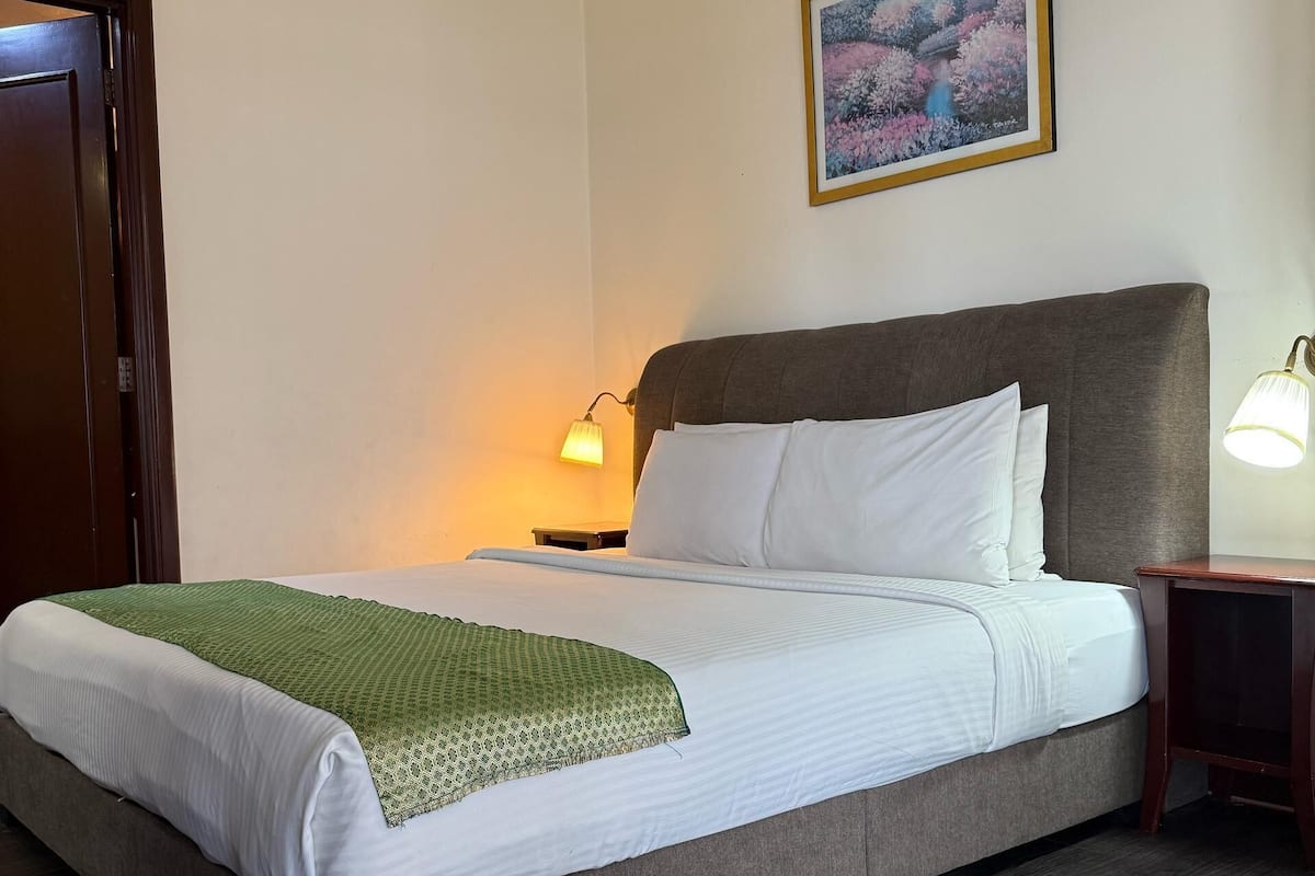 A neatly made bed with white linens and a green throw, a bedside lamp, a framed picture on the wall, and a wooden nightstand.