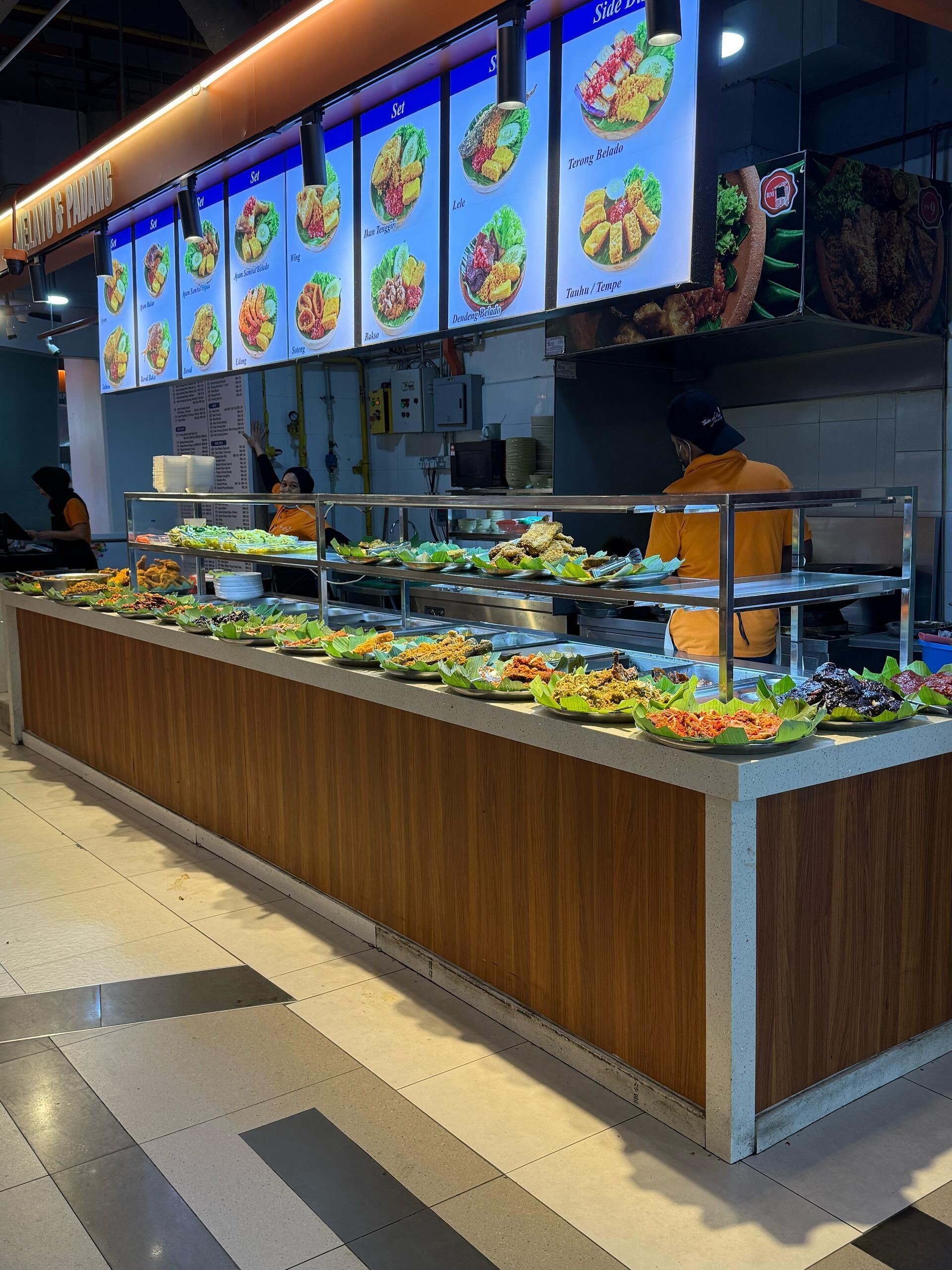 Un banco buffet con diversi cibi esposti su vassoi, un cartellone con il menu sopra e una persona che serve.