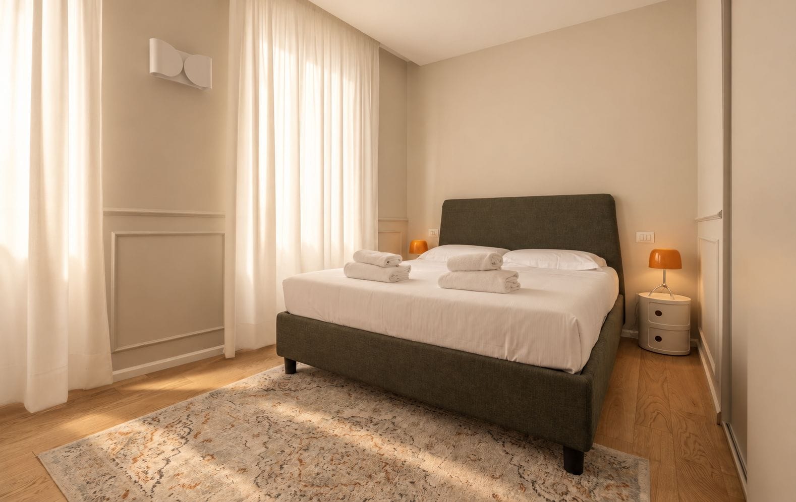 A neatly made bed with white linens and folded towels, a bedside table with a lamp, and a patterned rug on a wooden floor.