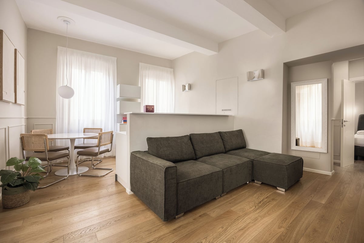 A modern living room with a grey sectional sofa, a dining table with chairs, and a wooden floor.