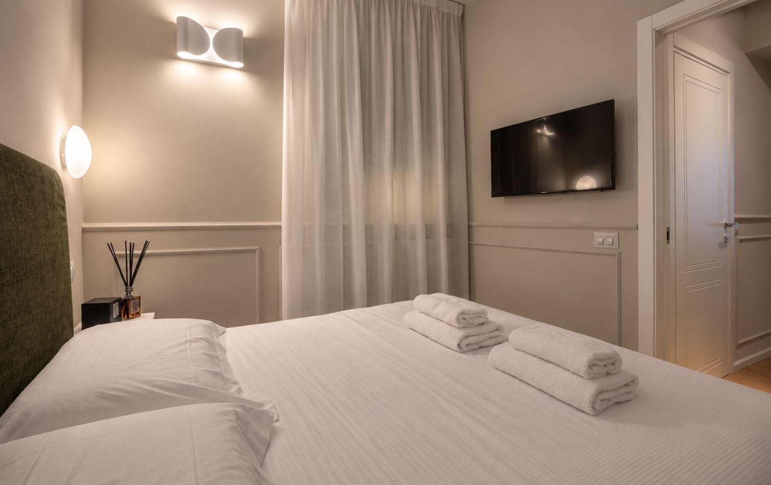 Quarto de hotel com uma cama, televisão de tela plana na parede, mesa de cabeceira com abajur e janela com cortinas.