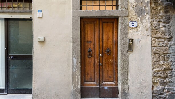Una puerta doble de madera con un tirador de metal en forma de anillo, un cartel con el número 2 y un buzón pequeño en una pared color beige.