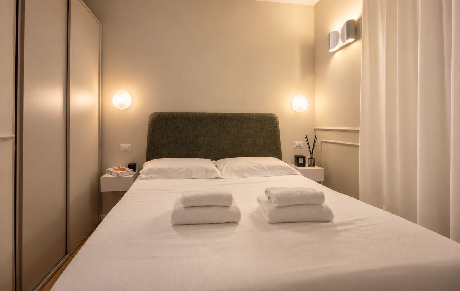 A neatly made bed with white linens and two folded towels, a bedside table with a lamp and a small box, and a wall-mounted light fixture.