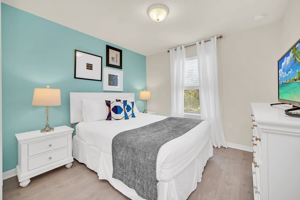 Une chambre à coucher comprenant un lit, des tables de chevet, une commode, un téléviseur et une fenêtre avec des rideaux.