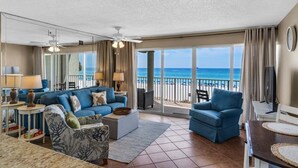 Un salon spacieux avec un grand canapé, des fauteuils et un téléviseur. Un balcon avec vue sur la plage et l’océan est visible.