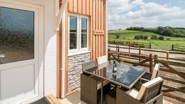 A room with a stone wall, a window with a nameplate, a glass table, wicker furniture, and a view of a green landscape. - Emminster (Weymouth)