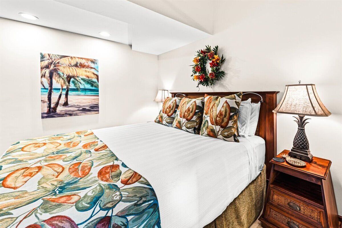 Une chambre à coucher avec un lit, une table de chevet, une lampe fixée au mur, un tableau représentant des palmiers et une guirlande accrochée au mur.