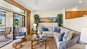 A modern living room with a glass coffee table, wicker chairs, a sofa with patterned cushions, and a large painting on the wall.