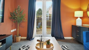 Een kamer met een schuifglasdeur, een potplant, een tafel met een vaas en kaars, een lamp en een televisie aan de muur.