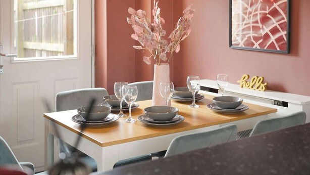 Een eettafel gedekt met borden, kommen en wijnglazen, een vaas met gedroogde bloemen, en een bordje met de tekst 'hello' aan de muur.