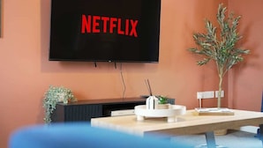 Een kamer met een flatscreen-tv aan de muur waarop 'NETFLIX' staat, een klein tafeltje met een wit voorwerp en een plant.