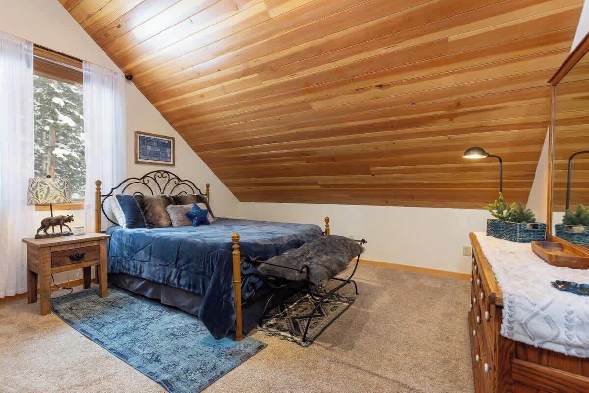 Une chambre avec un plafond en pente, un lit avec une tête de lit, une table de chevet, une lampe et une fenêtre avec des rideaux.