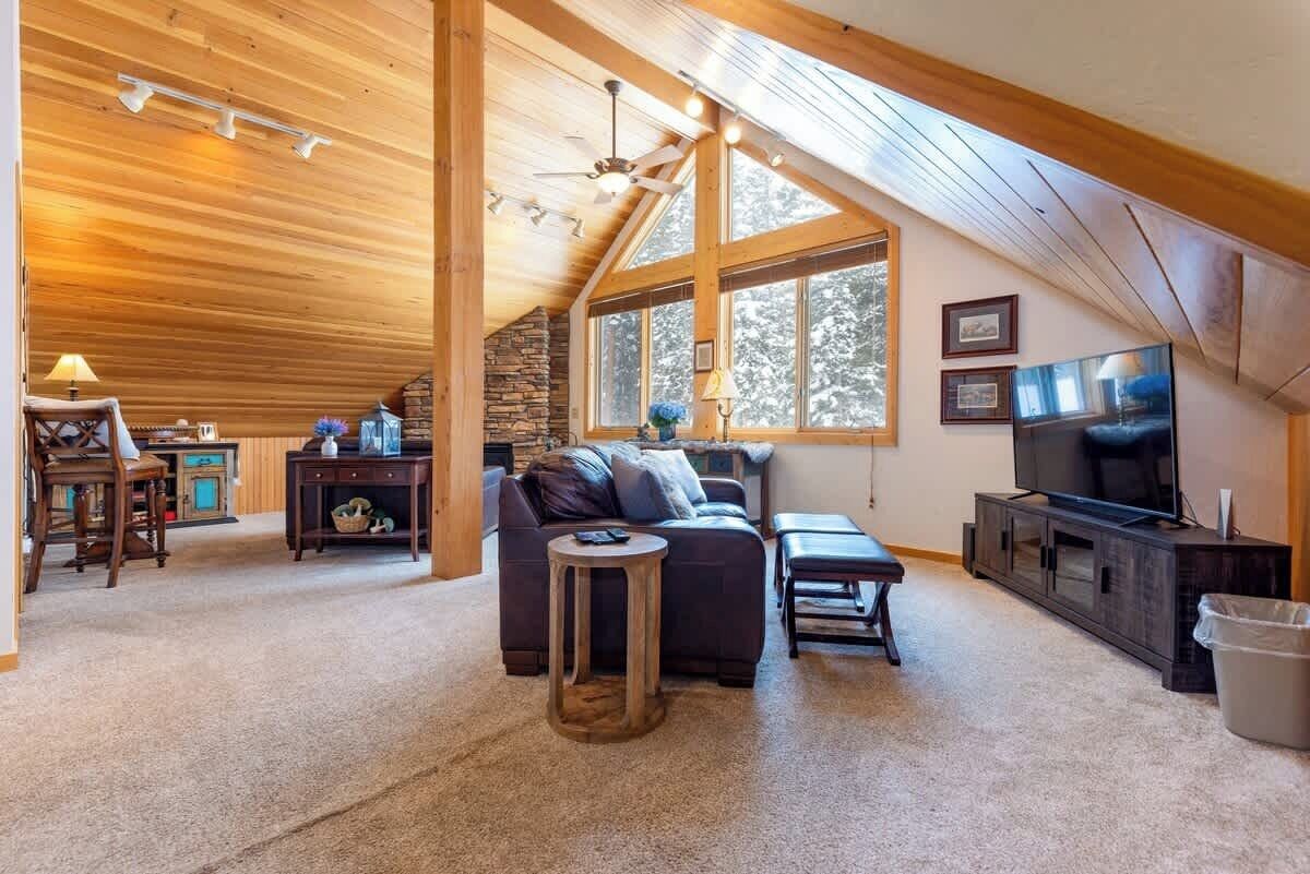 Ein gemütliches Wohnzimmer mit Holzbalkendecke, Kamin aus Stein, großem Fenster, einer Couch, einem Couchtisch und einem Fernseher.