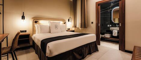 Habitación de hotel con cama, mesitas de noche, escritorio, silla, armario y baño con lavamanos y espejo.