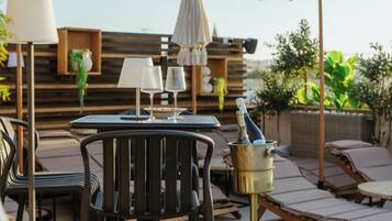 Un patio con una mesa dispuesta para dos personas, dos copas de vino, una botella en un cubo, sillones de descanso y un fondo de madera con plantas.