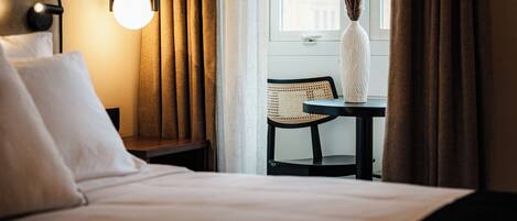A hotel room with a bed, a nightstand, a lamp, a window with curtains, a vase with dried plants, and a chair.